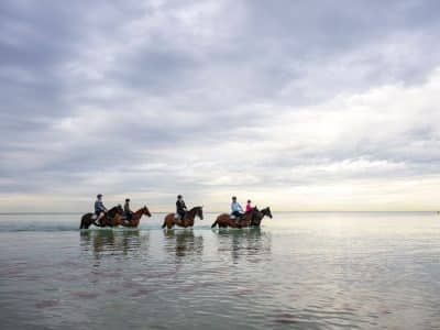 Horse riders wade through shallow water under a cloudy sky