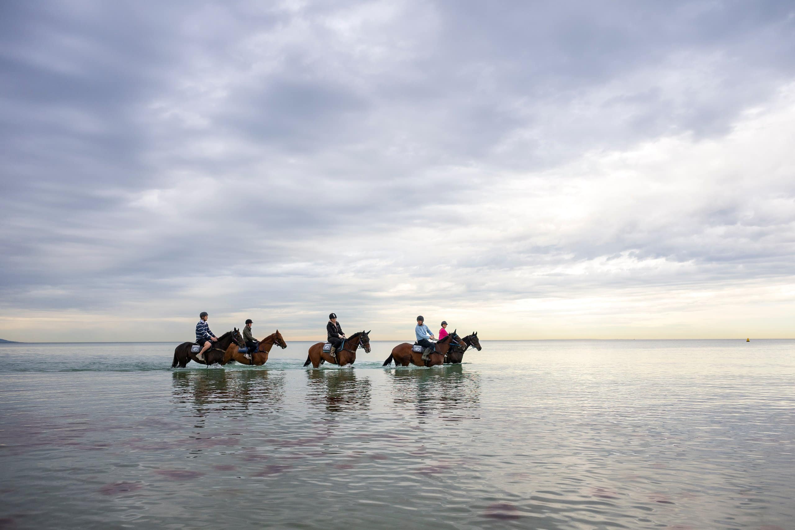 Four riders on horseback move through shallow seawater beneath a cloudy sky.
